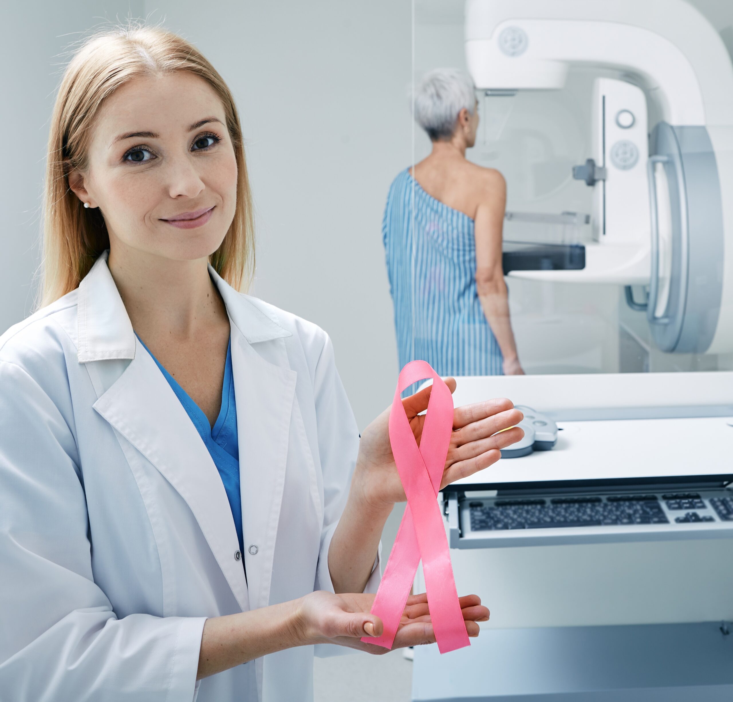Breast cancer ribbon held by medical professional with mammogram machine in background.